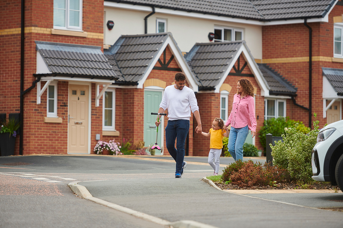 A man and a woman holding hands with a child in front of a row of homes. The man holds a scooter.