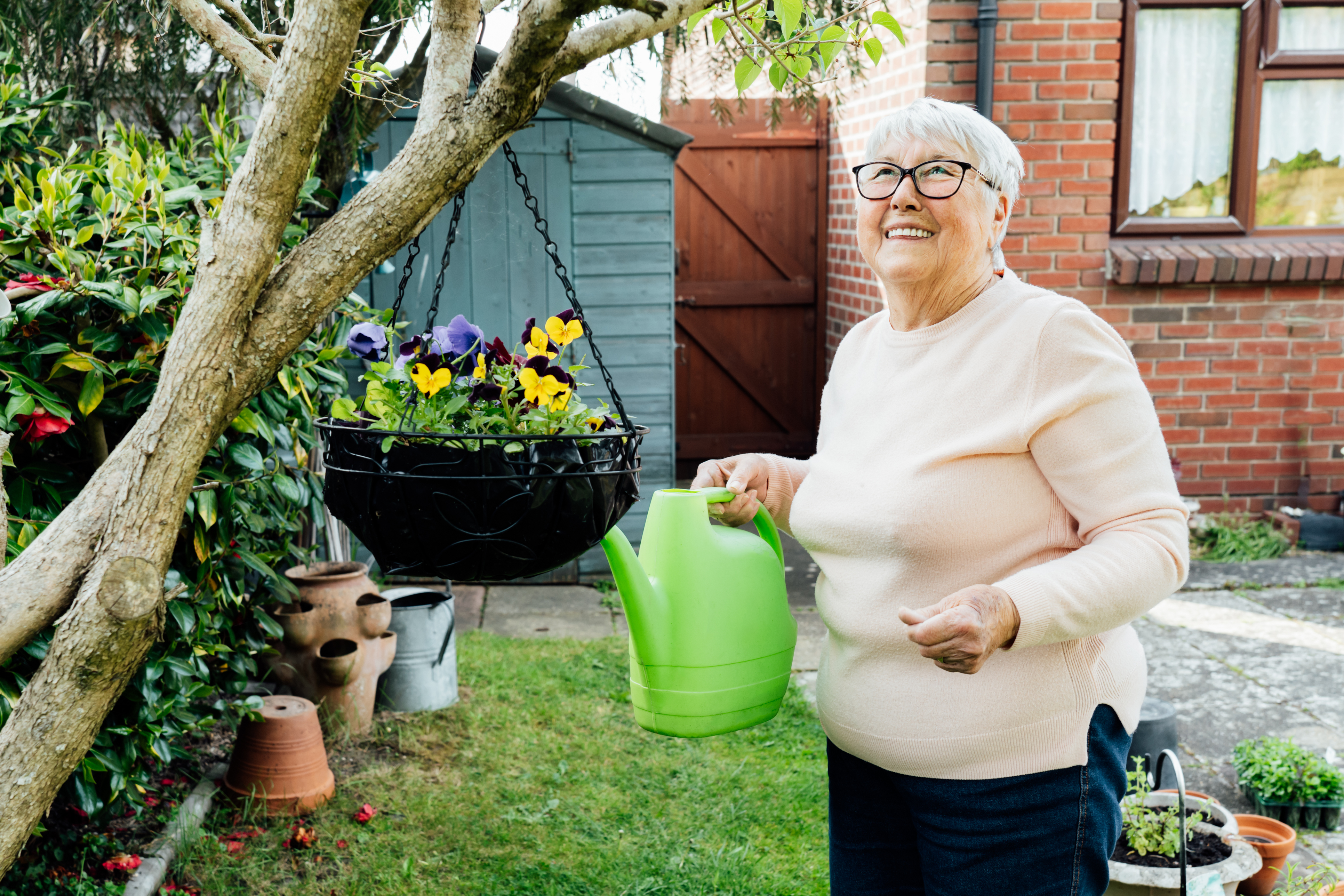 Older woman in her garden watering hanging basket with green watering can, smiling, looking upward