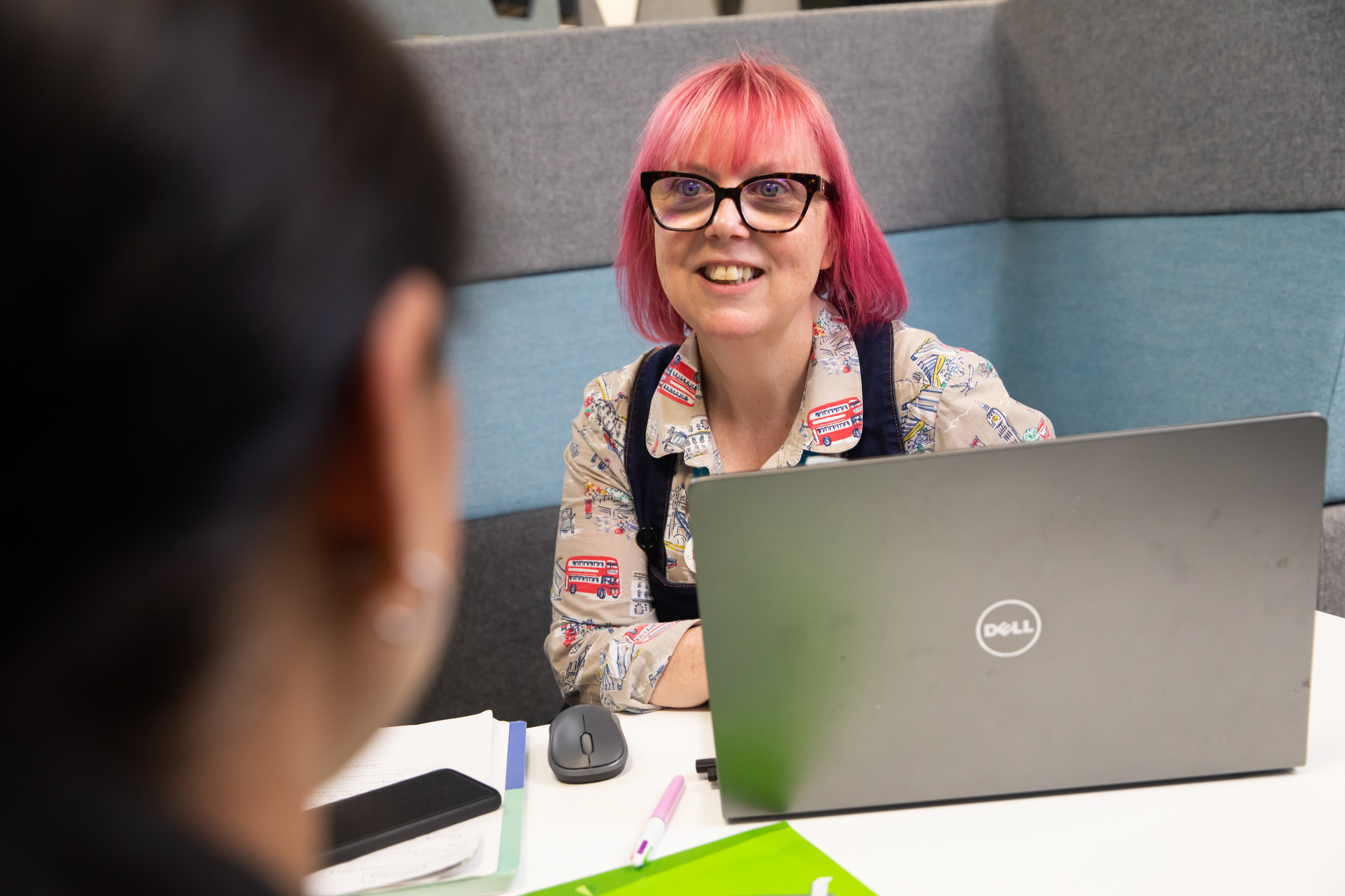Colleague with pink hair on laptop, sat facing another colleague who is blurred in foreground