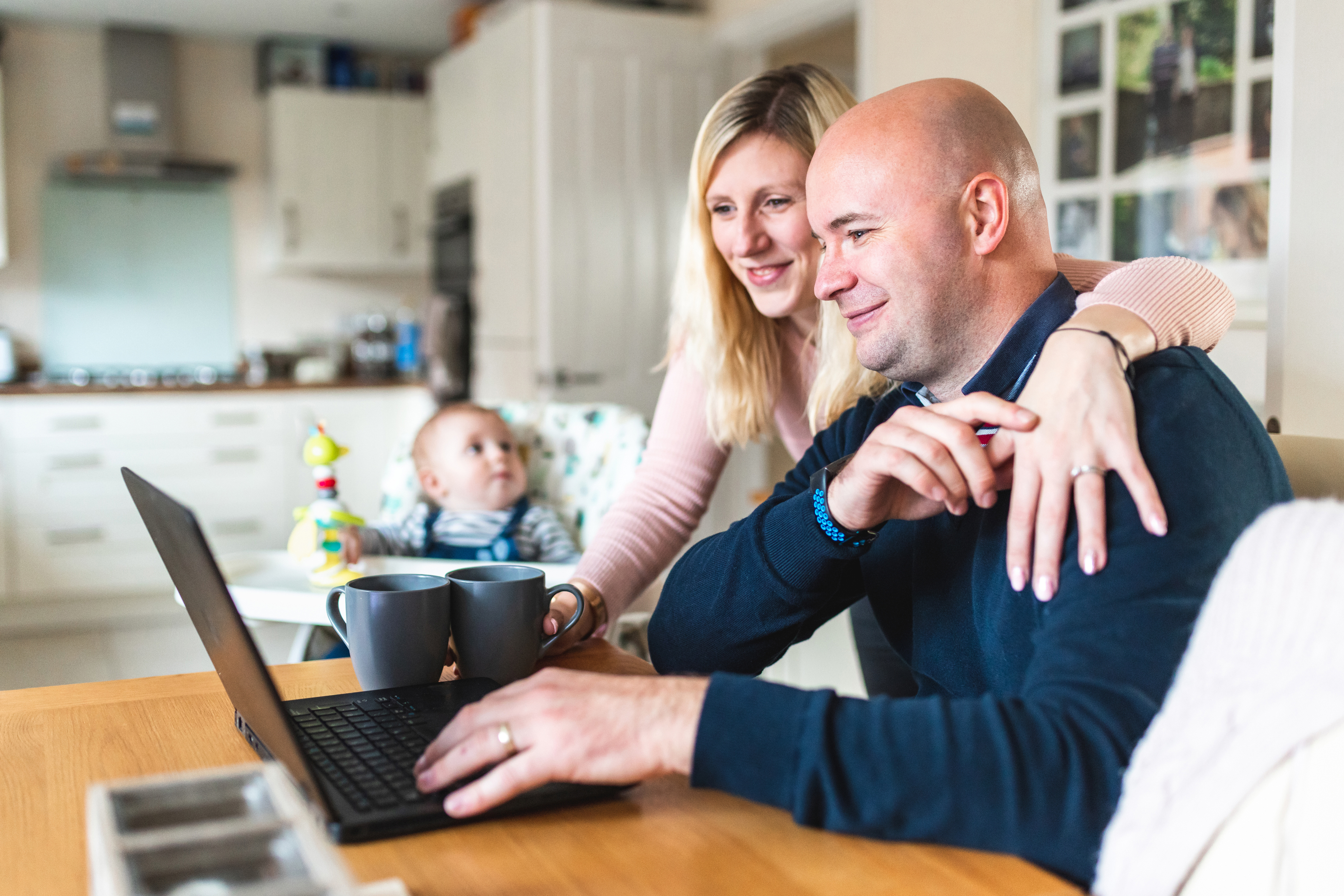 Mother and father looking at laptop on kitchen table, baby in high-chair in the background