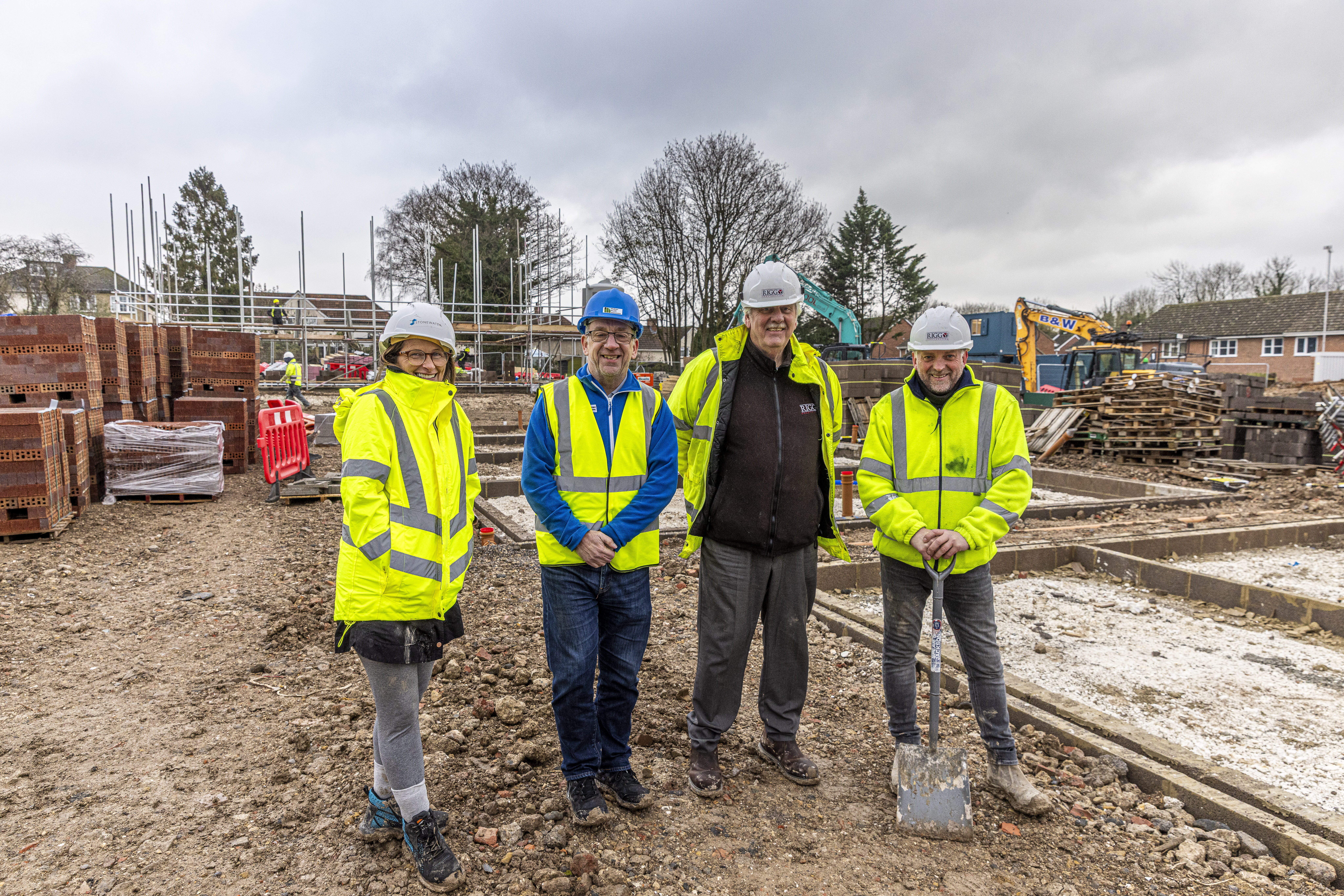 Four people stand in high vis vest and hardhats on a building site, one leans on a spade