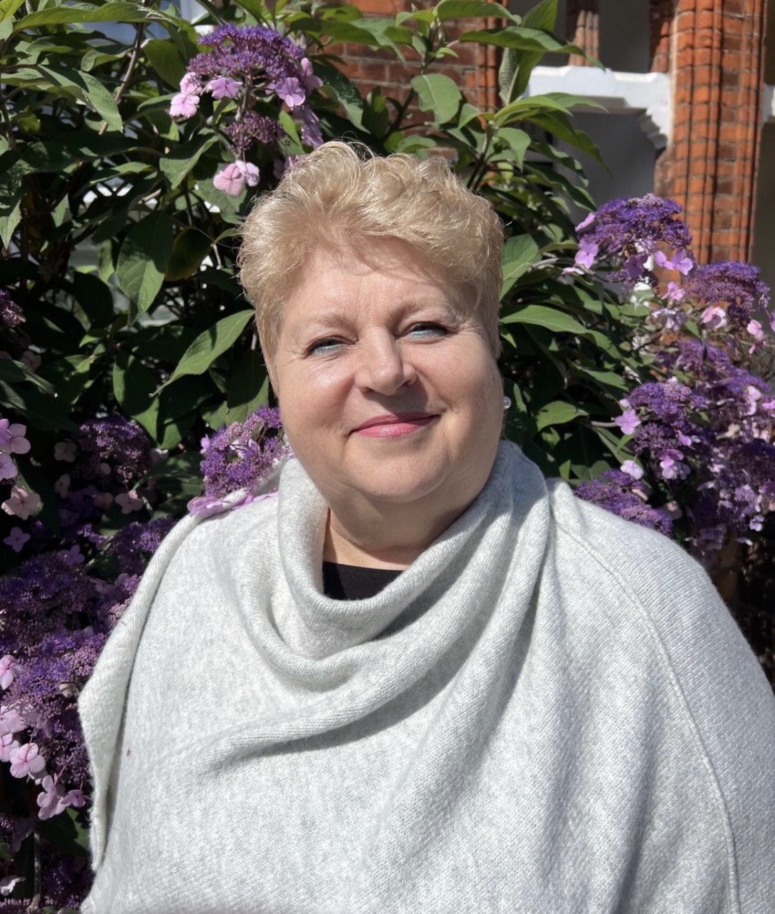 Lady smiles at camera, she is wearing a grey jumper and has short curly blonde hair.