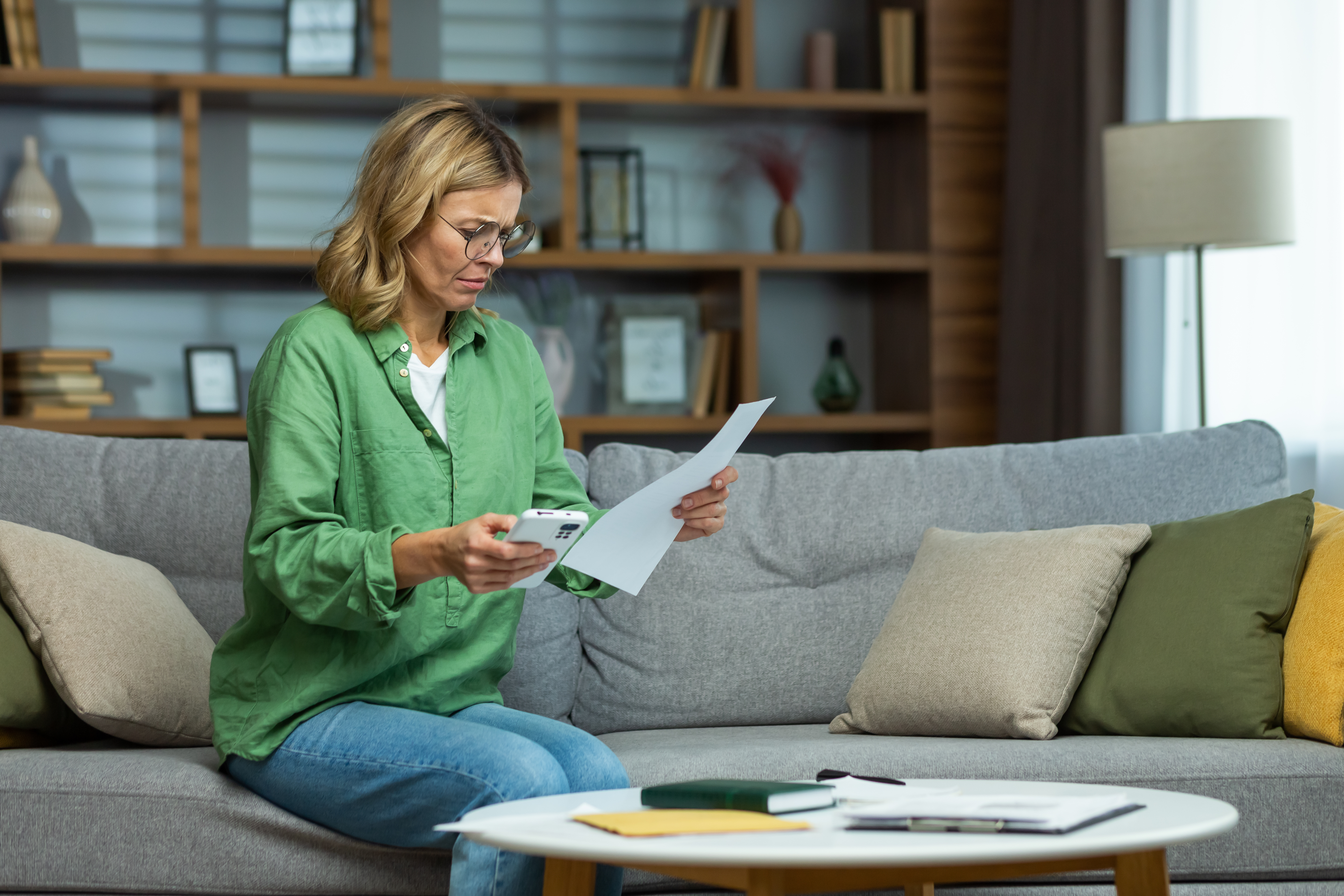 Blonde woman on sofa looking at paperwork in her hand with concerned expression