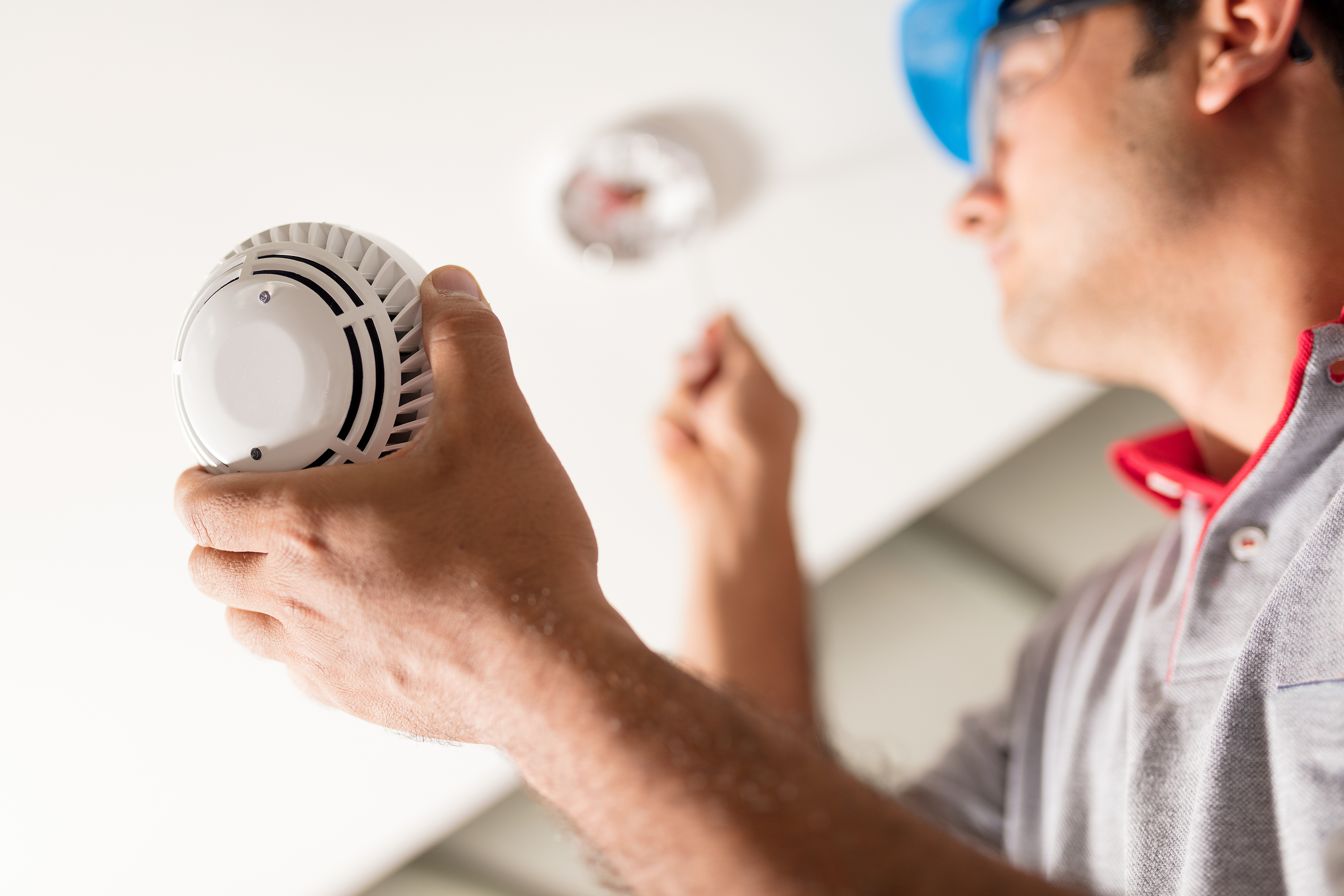 Close-up of a tradesperson in a blue hard hat replacing the battery in a ceiling fire alarm