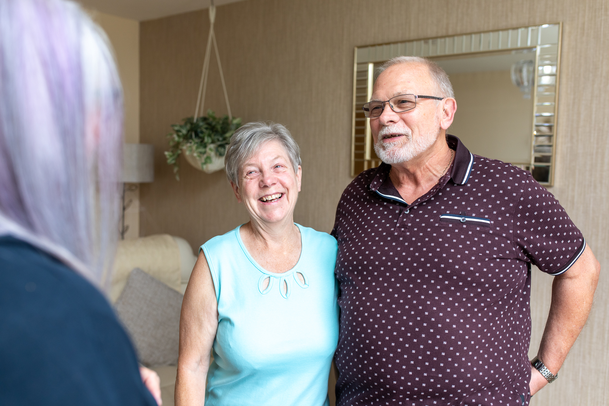 Older couple in their home facing camera, smiling and talking to person with long mauve hair