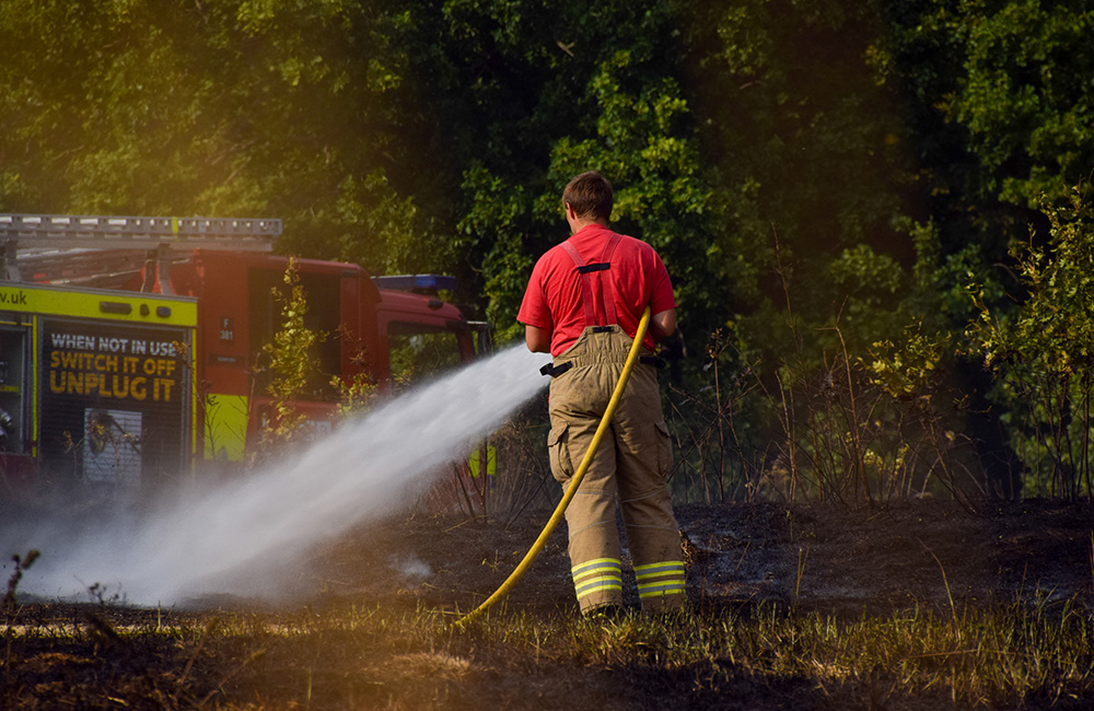 Fireman stood spraying fire with a hose. A fire engine is in the background.
