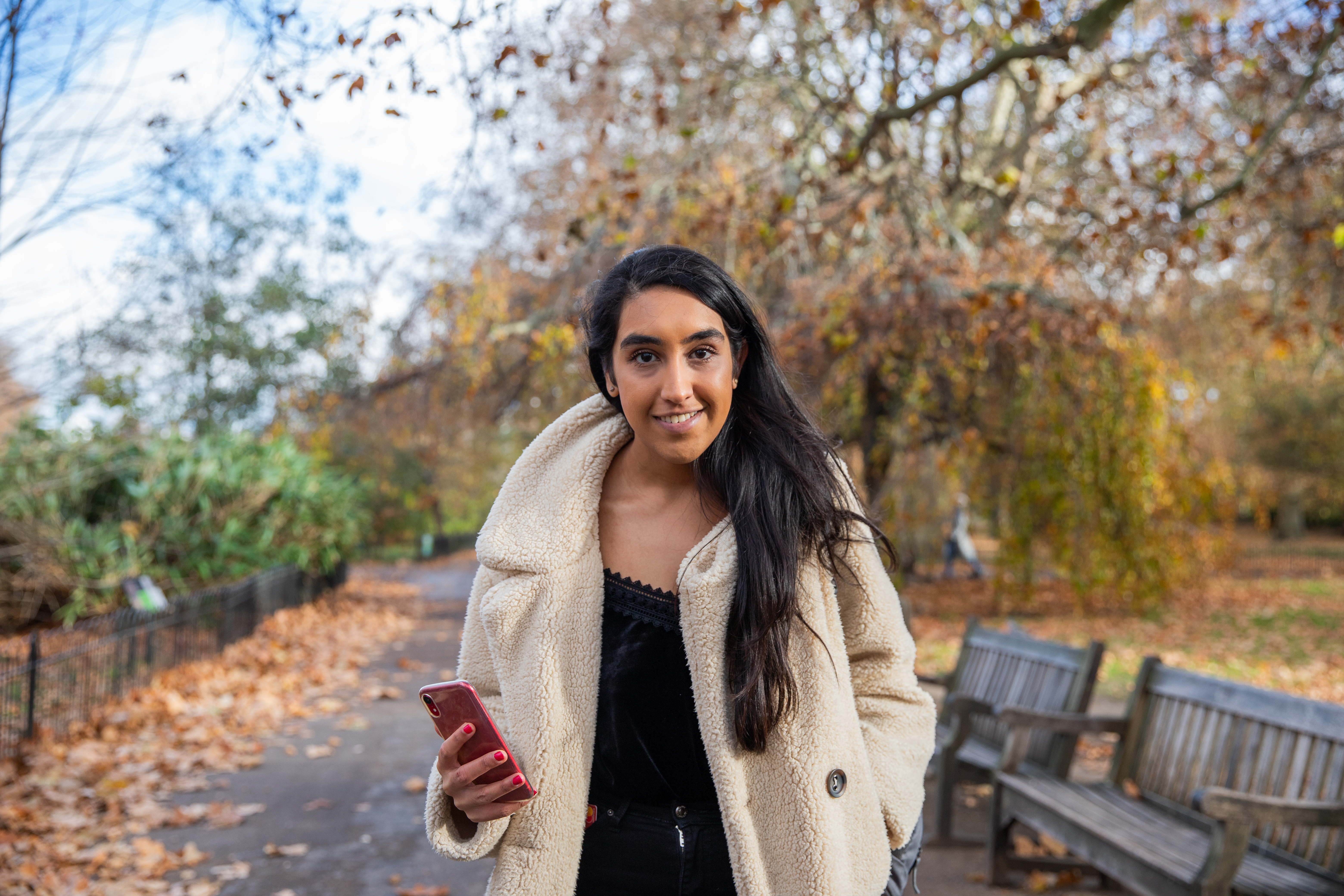 Smiling woman stood outside under autumn trees holding mobile phone