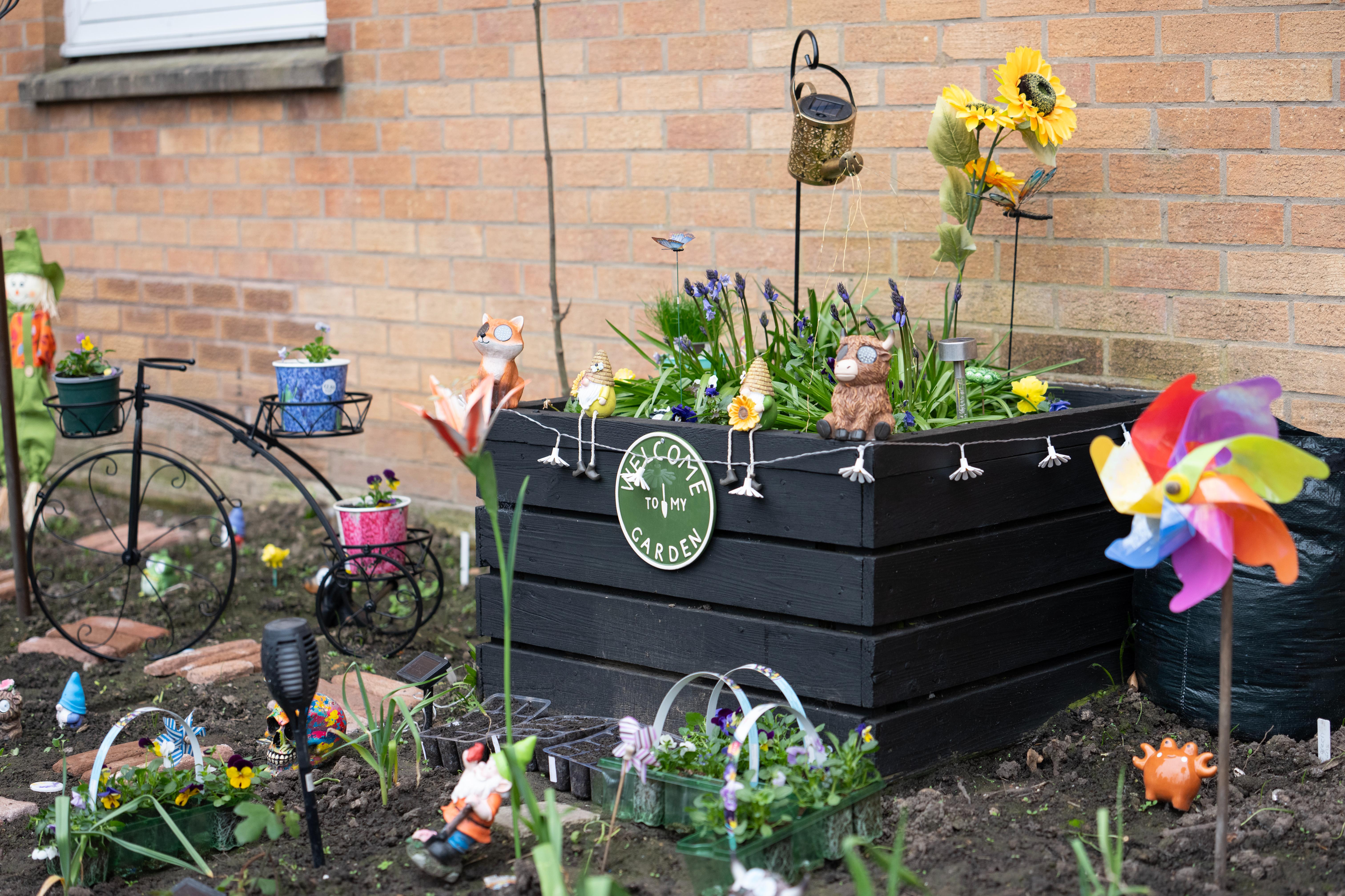 a black raised flower bed sits against a brick wall with seedlings and garden ornaments round it