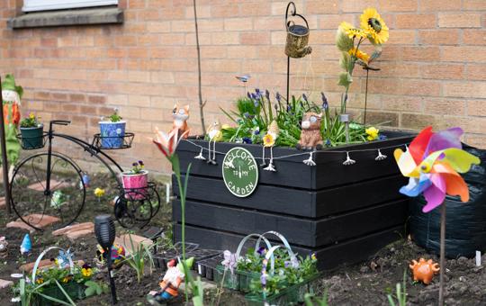 Black raised flower bed sits against a brick wall with seedlings and garden ornaments round it