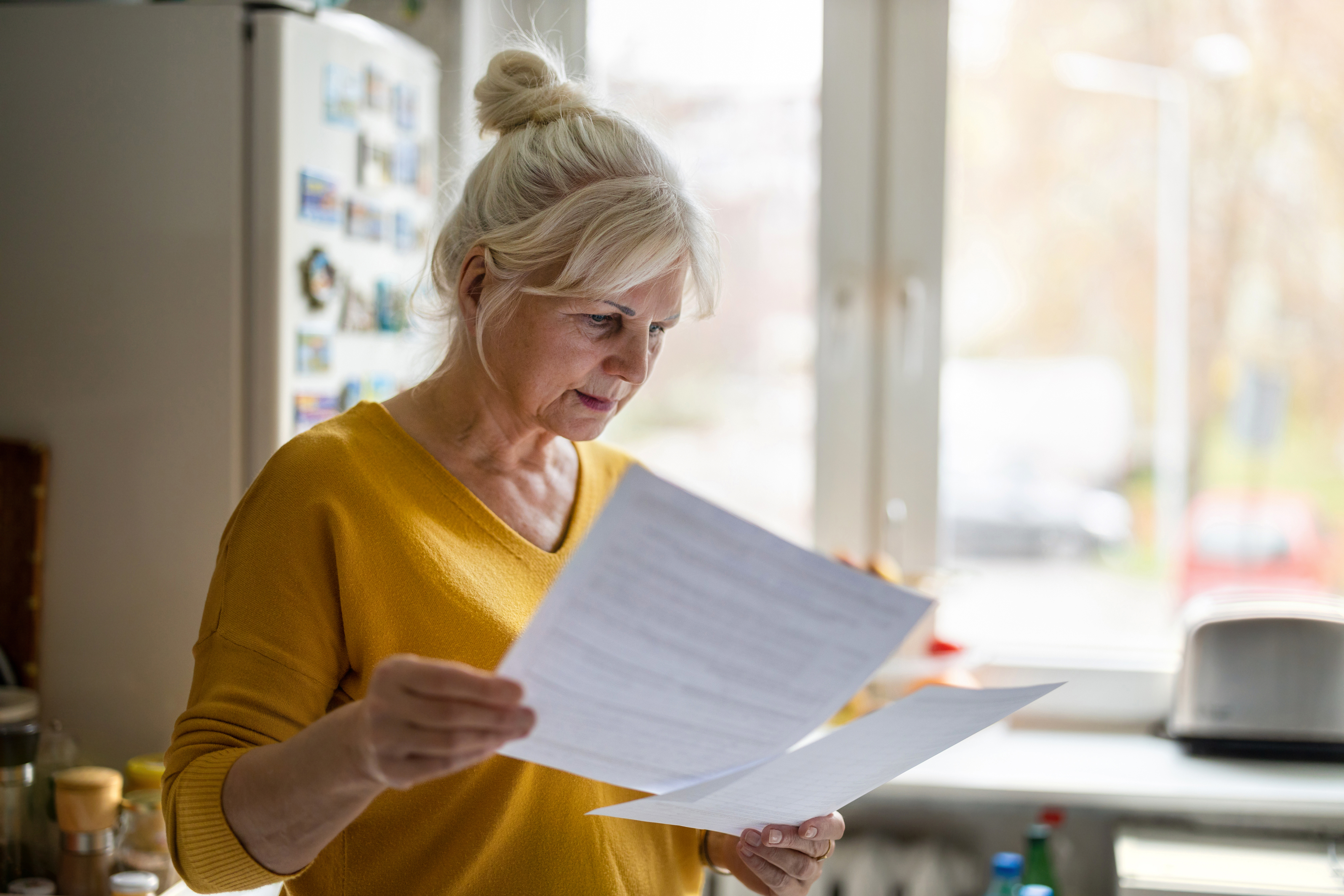 Woman in yellow top stood in kitchen, holding two pieces of paper with neutral expression