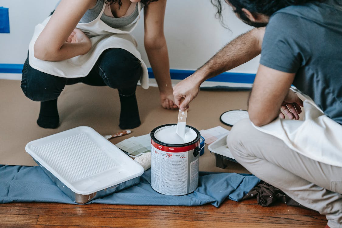 Man and woman crouched on wooden floor with dust sheets, white paint pot, and paint tray