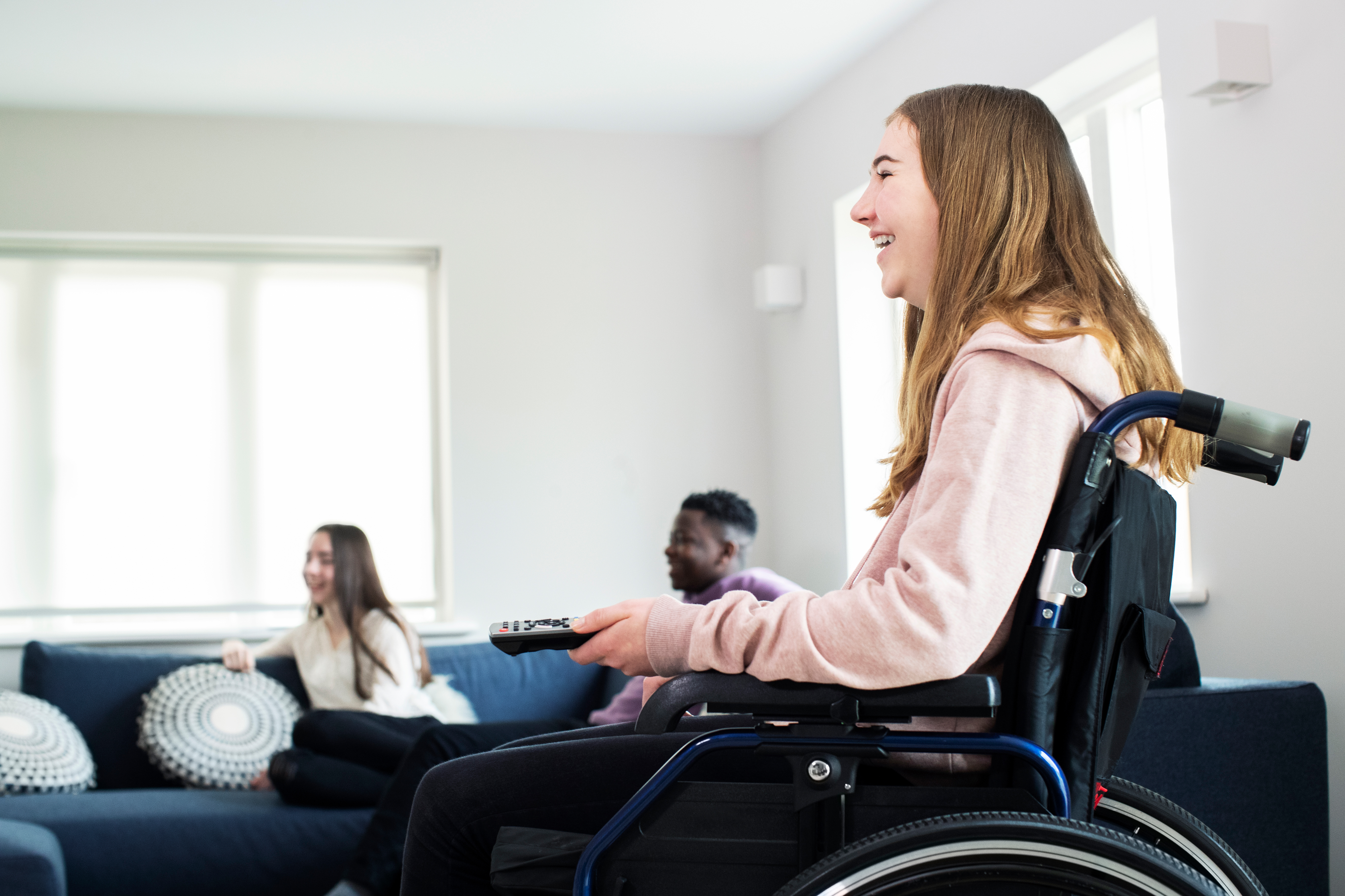 Young woman in wheelchair holding TV remote, friends sat on sofa in background