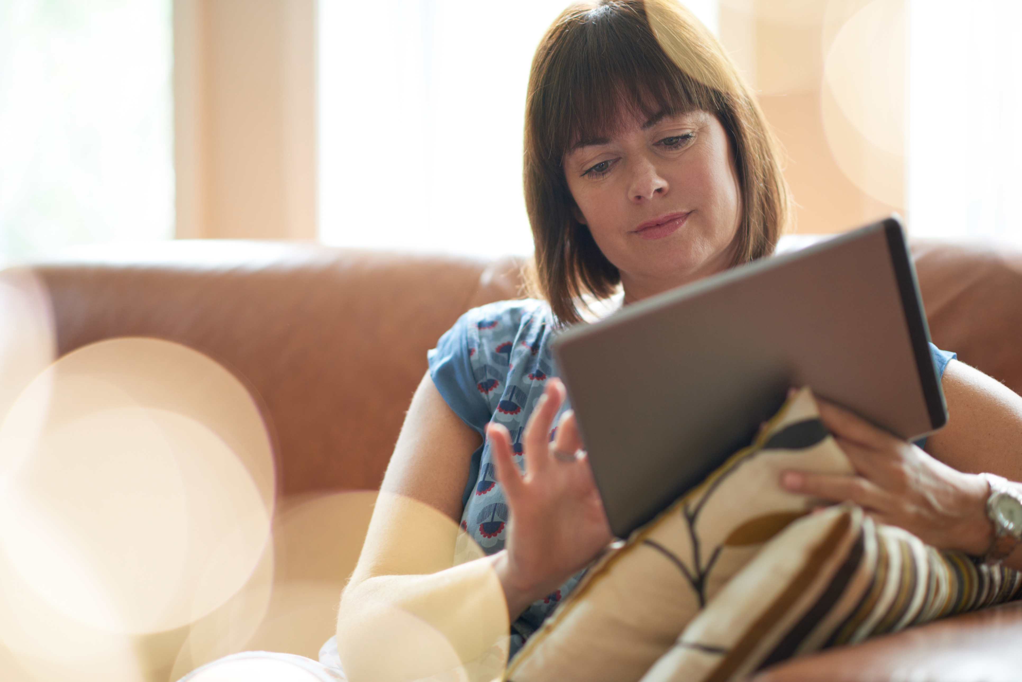 Woman looking relaxed, sat on sofa browsing on tablet device. Bokeh effect in foreground