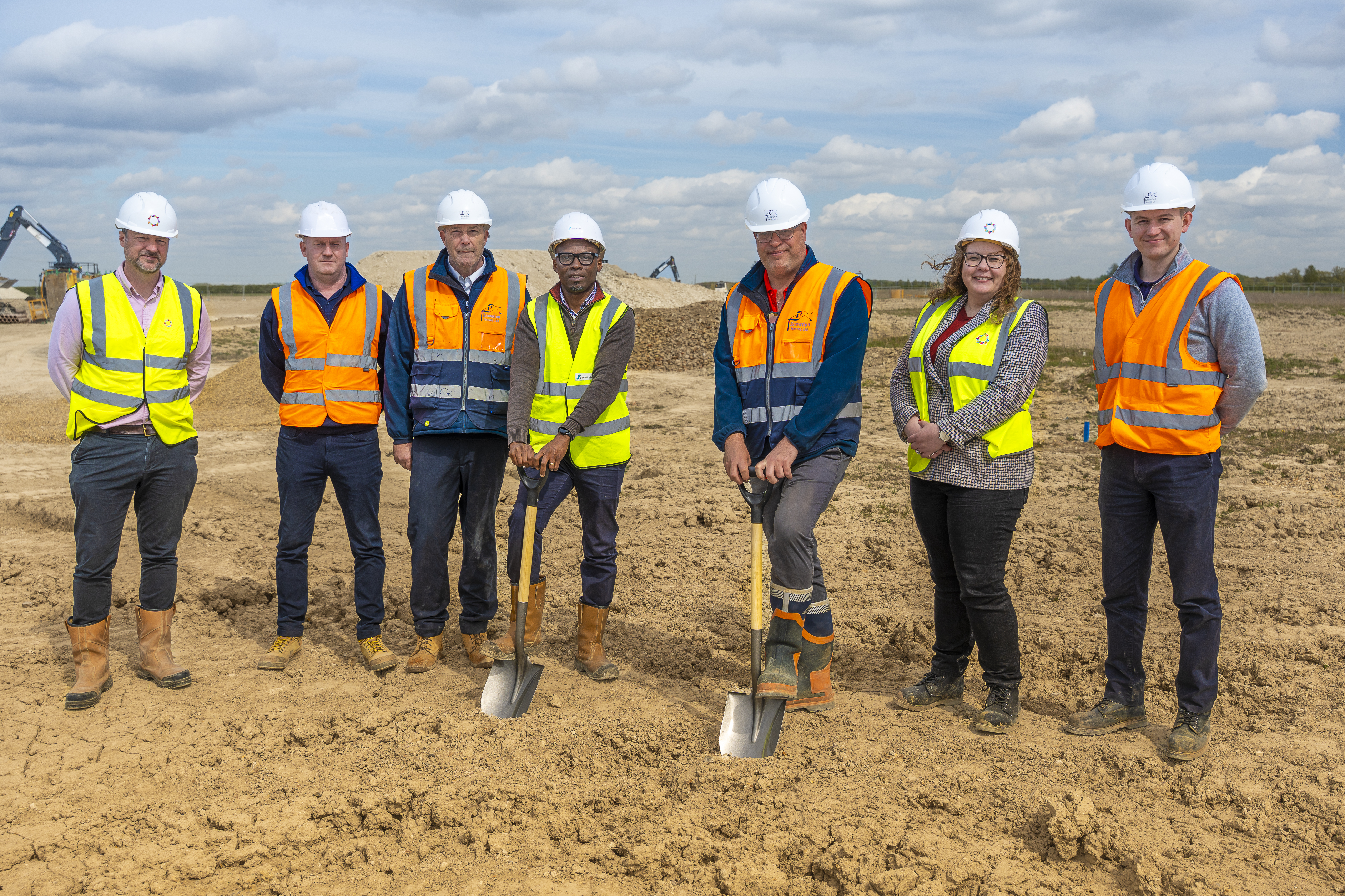 7 people in a row wear PPE boots, high vis and hats. Two are leaning on spades.
