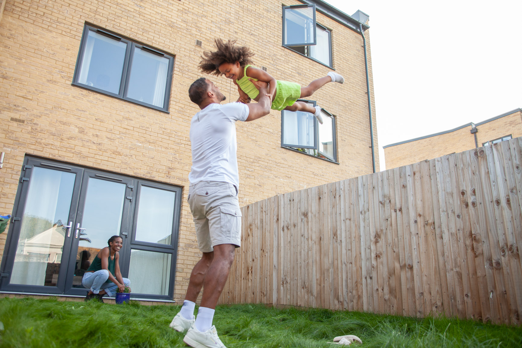 Young family in rear garden. Father is swinging daughter above his head.