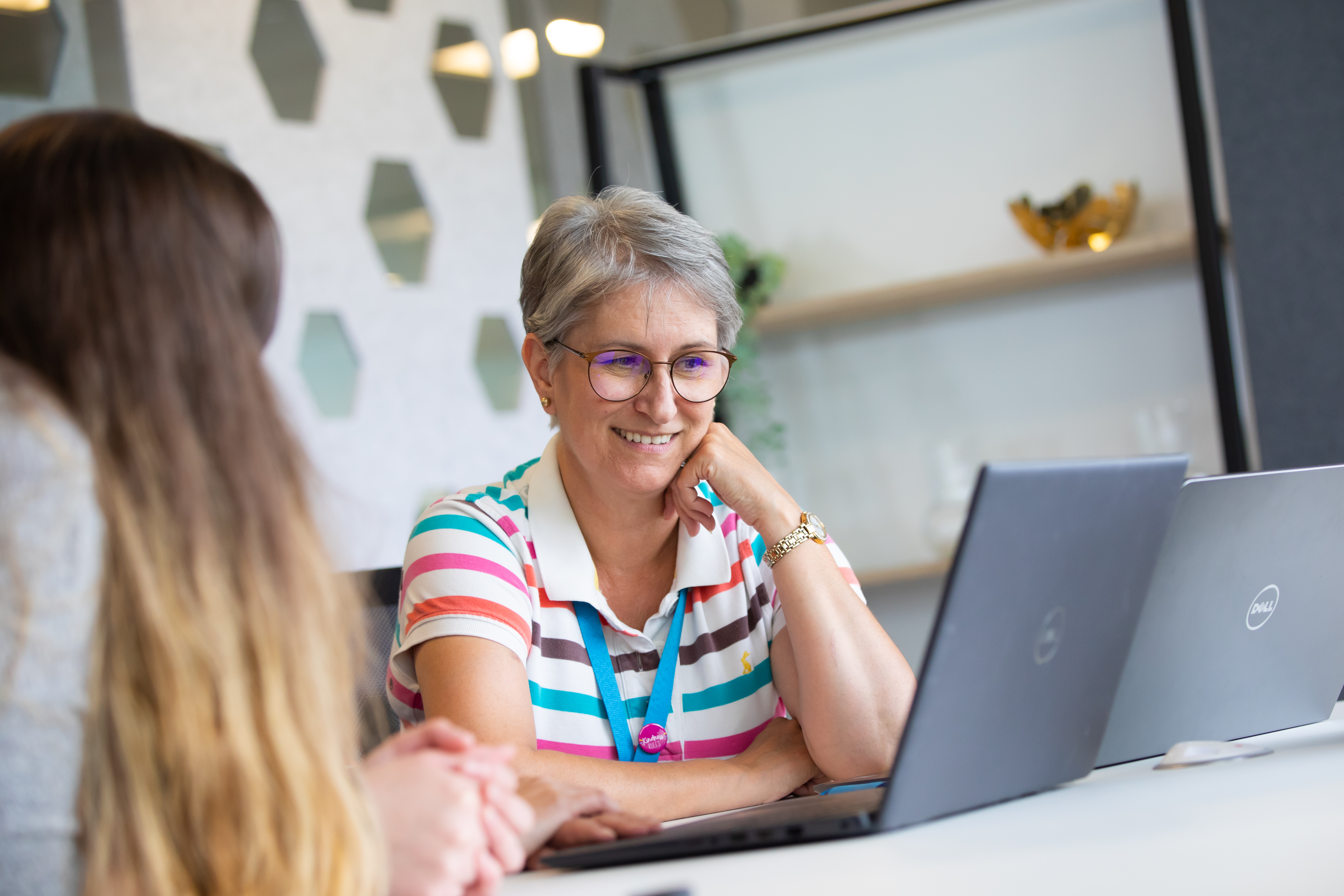 Smiling female colleague looking at laptop, sat next to female colleague blurred in foreground