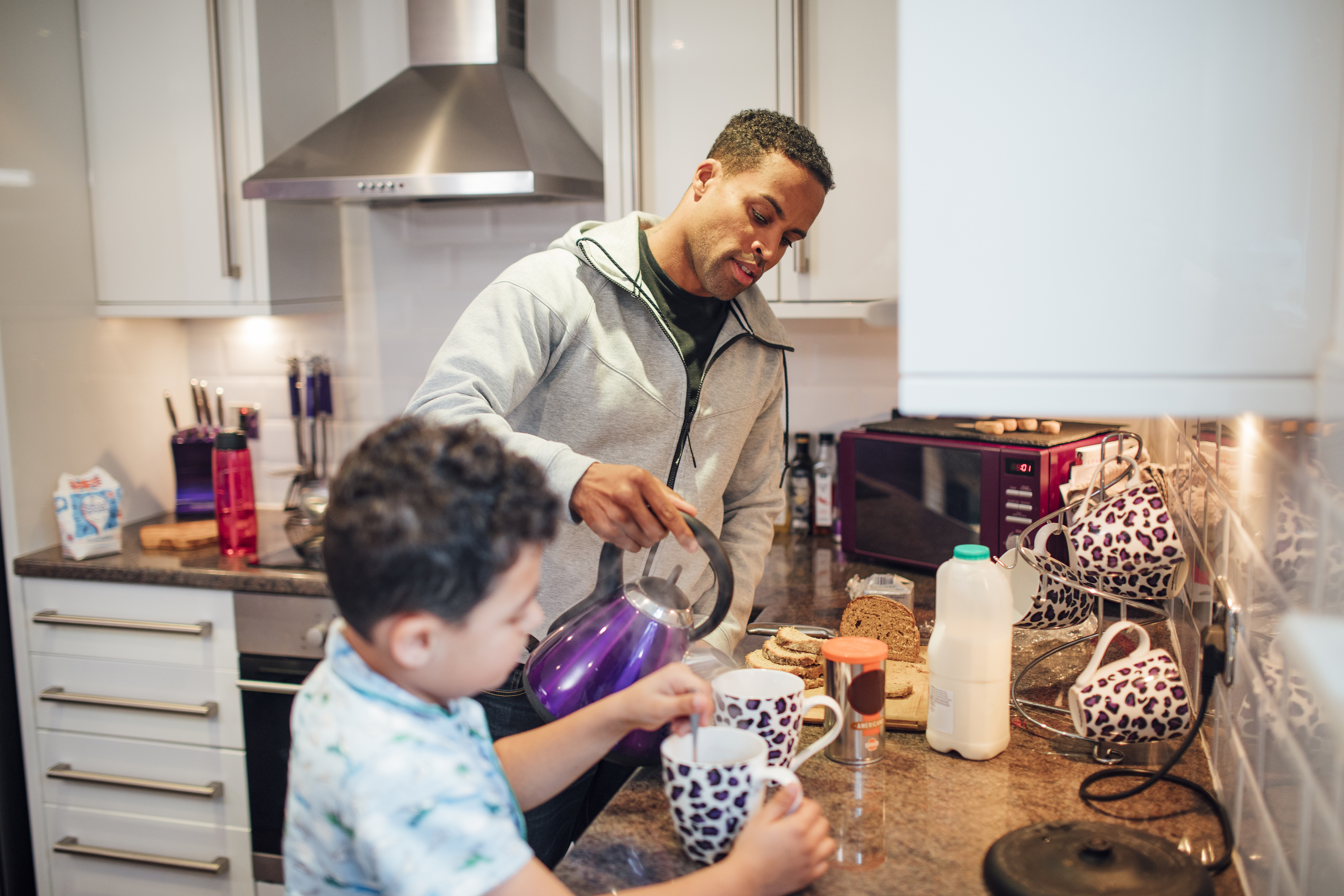 Young father with son making breakfast in kitchen. Son is stirring tea, father is pouring kettle.