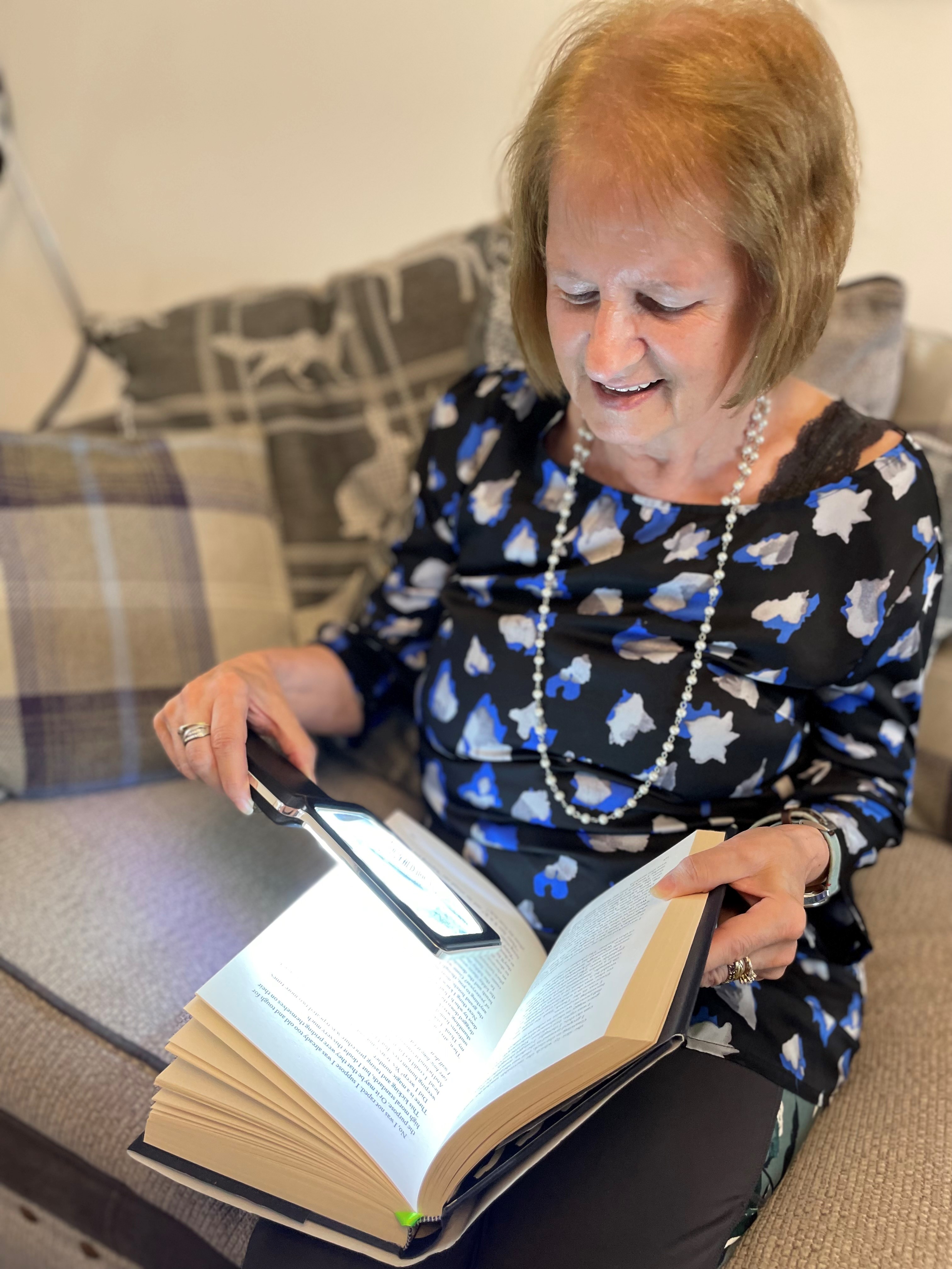 Older resident sat on chair using magnifying glass with light to read her book 