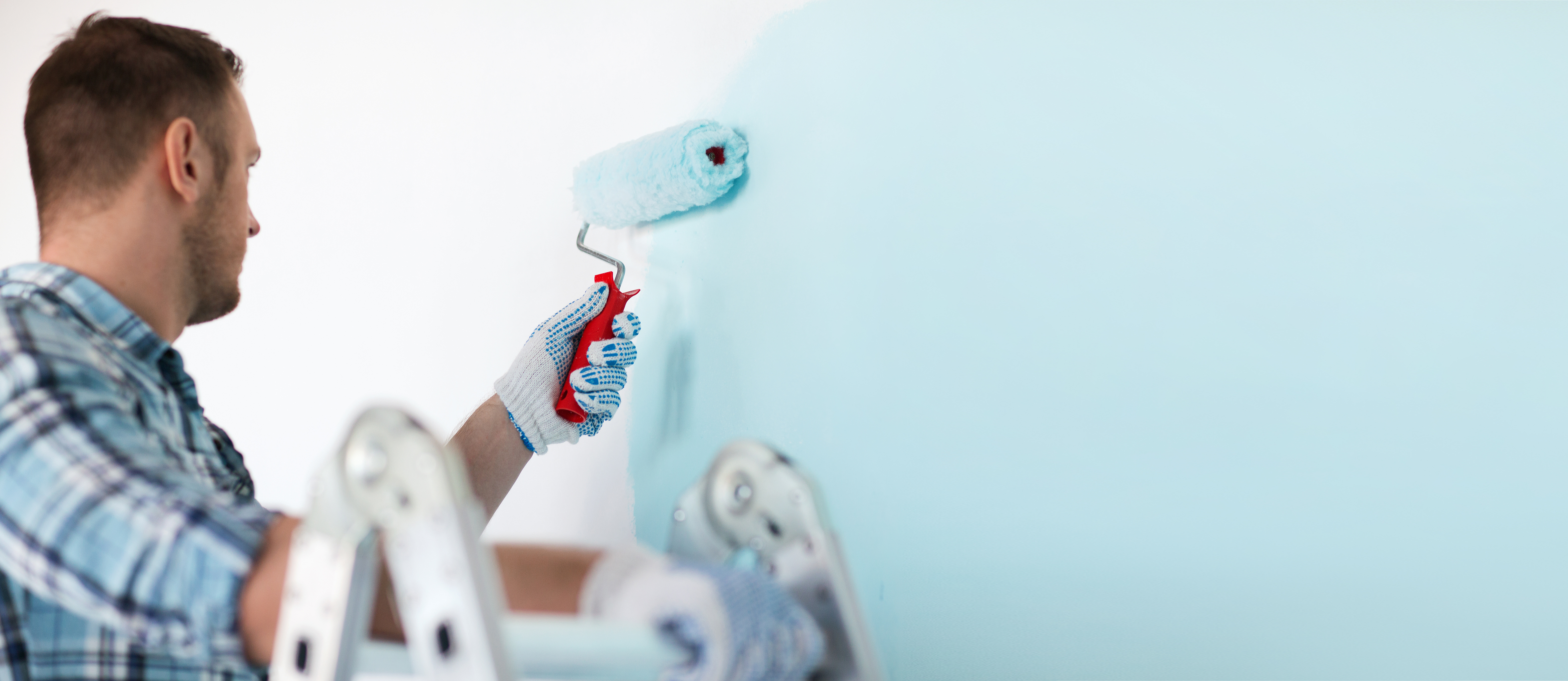 Man in blue checked shirt and gloves using roller to paint a baby-blue wall