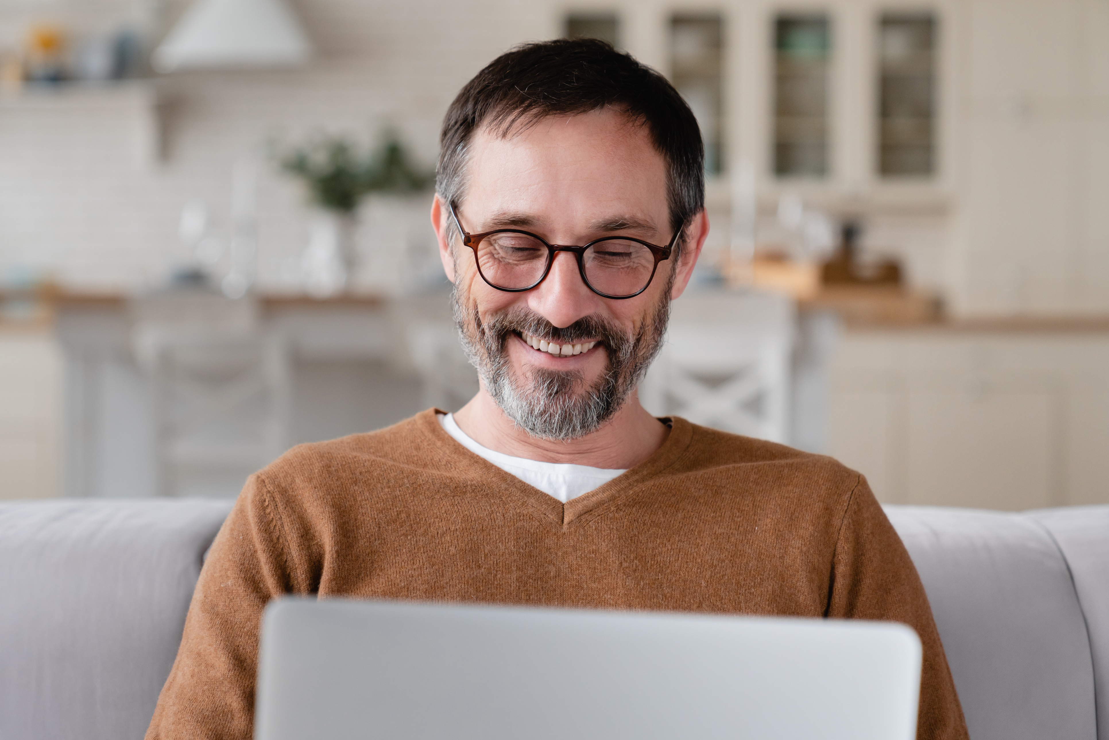 Man with glasses sat facing camera, smiling and looking down at laptop on his lap