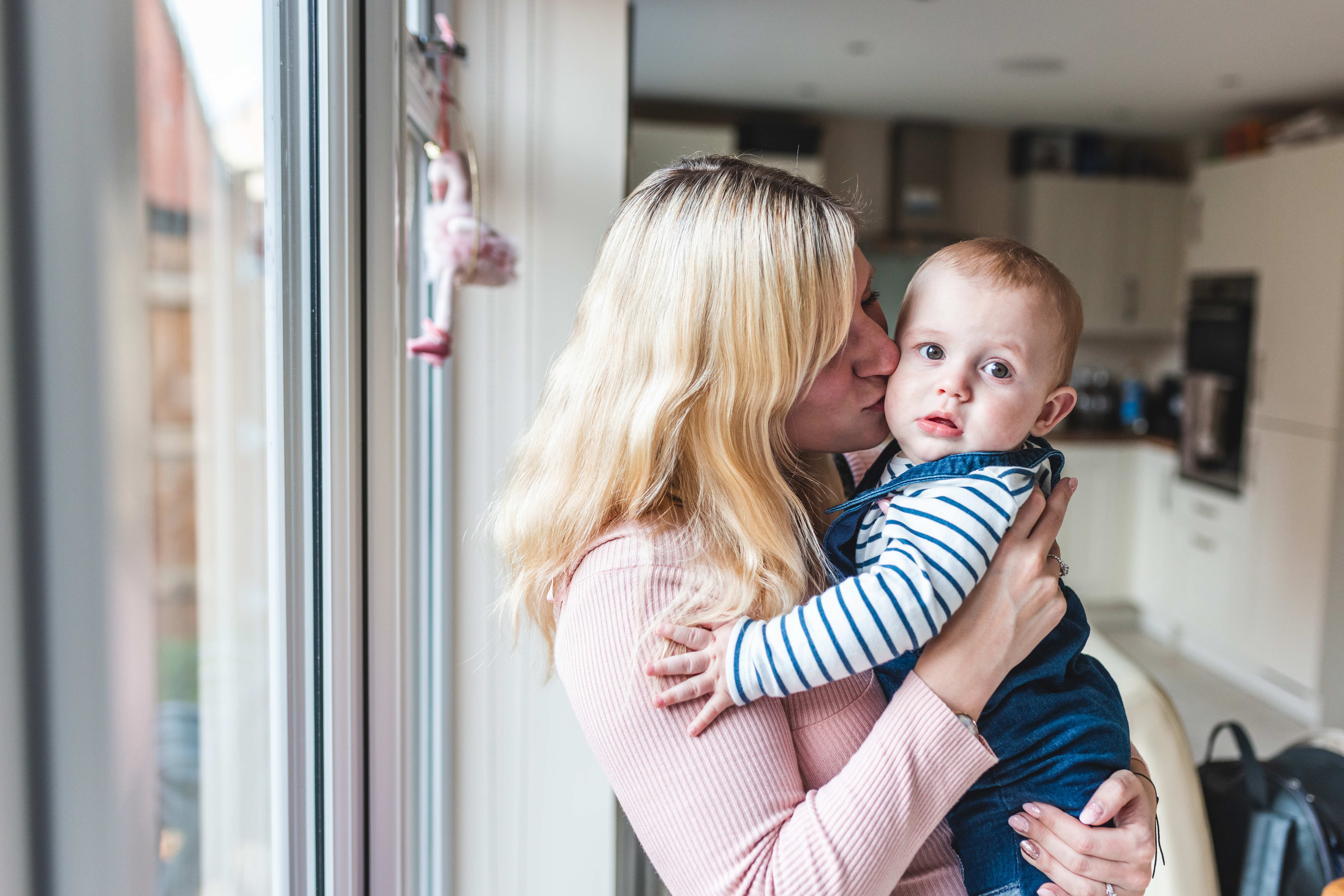 Blonde mother in pink jumper stood in kitchen near patio door, holding baby who is looking at camera